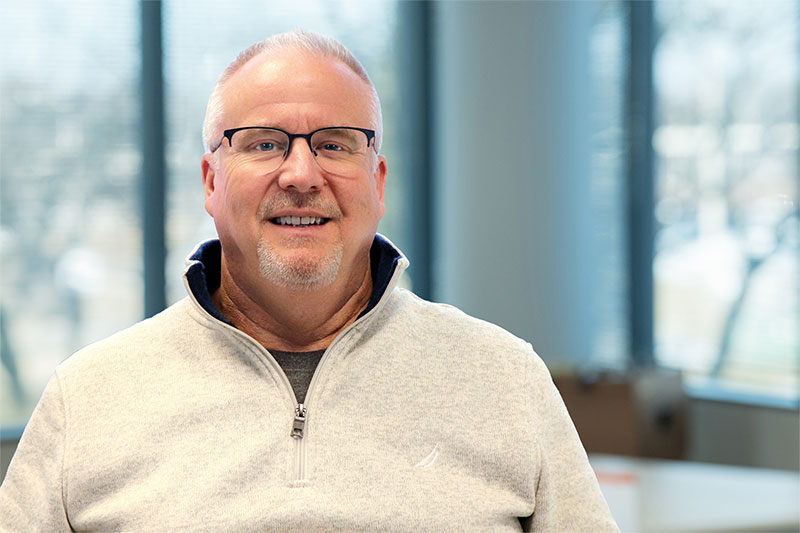 Smiling man with short gray hair and glasses wearing a light-colored quarter-zip sweater, standing in a bright office with large windows behind him.