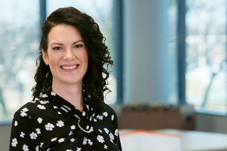 Smiling woman with dark curly hair wearing a black patterned blouse, standing in a bright office with large windows and a softly blurred background.