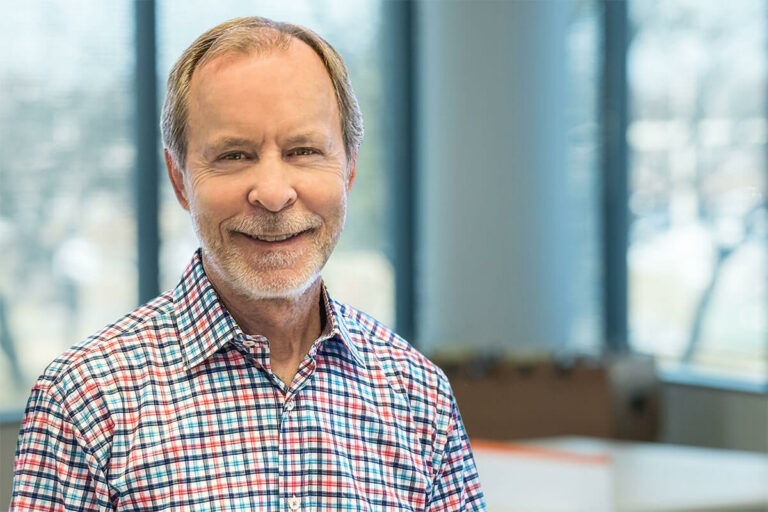 Smiling man with short gray hair and light beard wearing a colorful checkered button-up shirt, standing in a bright office with large windows behind him.