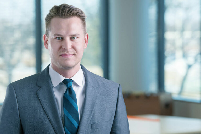 Man wearing a gray suit and blue striped tie stands in a bright office, looking at the camera with large windows and a blurred workspace behind him.