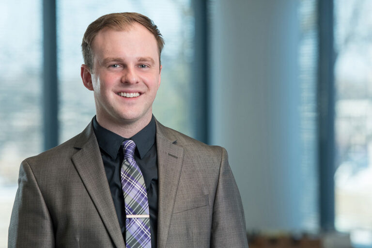 Smiling man wearing a brown suit jacket, black shirt, and patterned tie stands in a bright office with large windows and a softly blurred background.