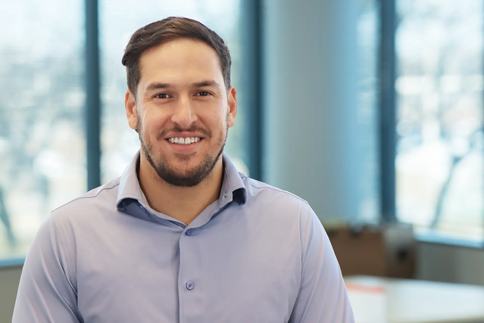 Smiling man with short dark hair and trimmed beard in light blue shirt, office background
