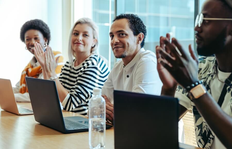 Diverse coworkers seated at table with laptops, clapping during meeting