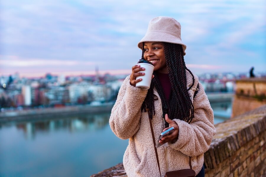 Woman in bucket hat sipping coffee by riverside with city skyline at dusk