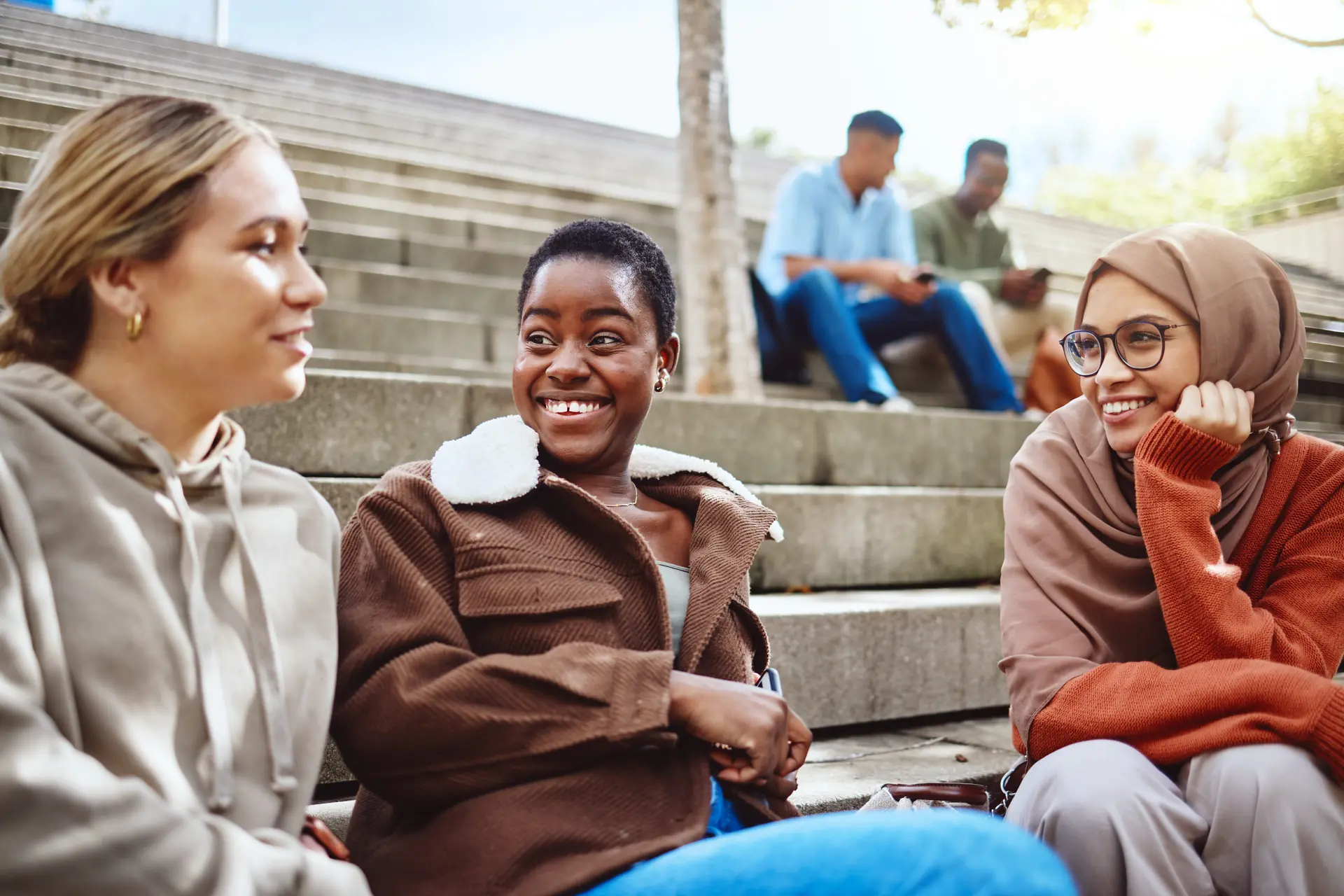 Three diverse women smiling and chatting on outdoor steps, two men behind them