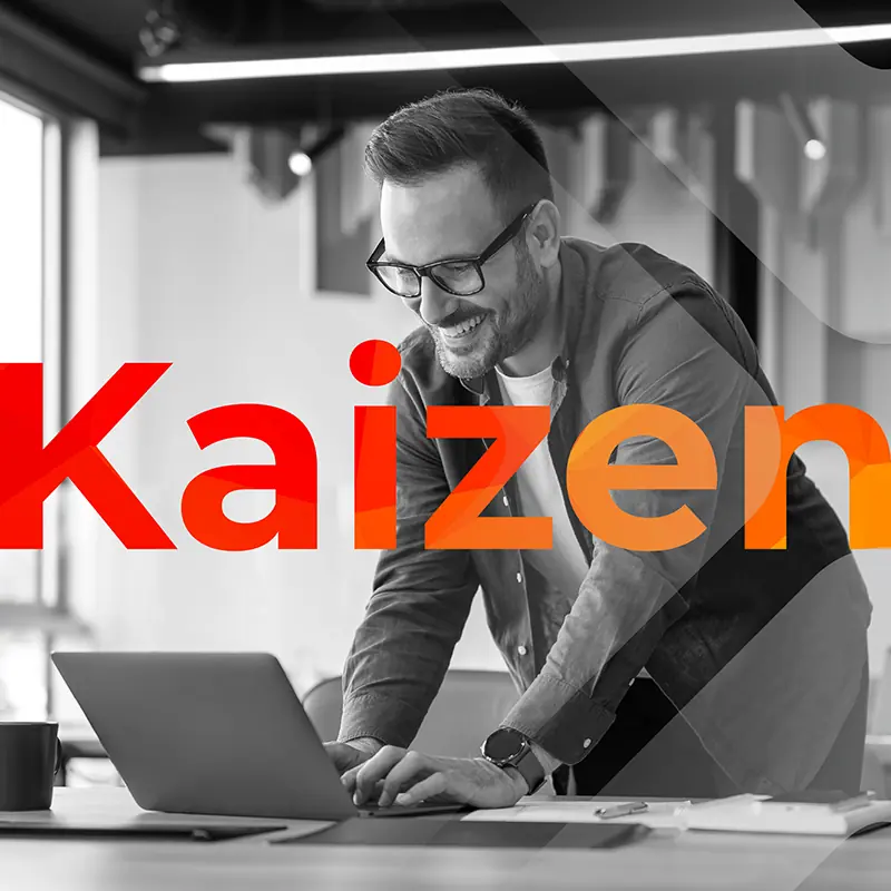 Smiling man working at a laptop with ‘Kaizen’ text overlay A man wearing glasses leans over a desk smiling while typing on a laptop, with papers nearby and bold ‘Kaizen’ text layered over the image.