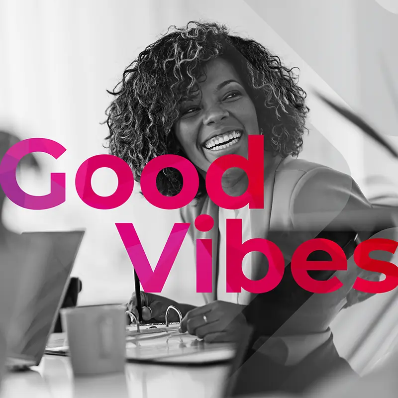 Smiling woman working at a desk with ‘Good Vibes’ text overlay A smiling woman with curly hair sits at a desk writing in a notebook, with laptops nearby and bold ‘Good Vibes’ text layered over the image.