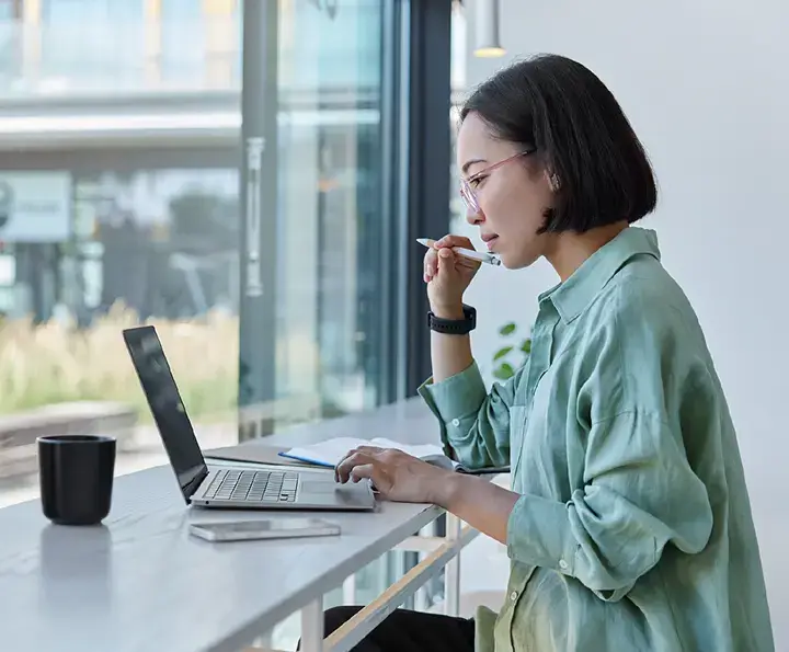 Woman in green shirt working on laptop at window desk with coffee mug